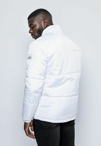 Vit pufferjacka med hög krage, med horisontell sömnad och en logotyp på vänster ärm. Bärs med svarta jeans.