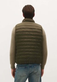 Vesta tăiată, de culoare verde măslin, cu un design matlasat orizontal, guler înalt, purtată împreună cu un pulover tricotat bej și blugi din denim albastru.