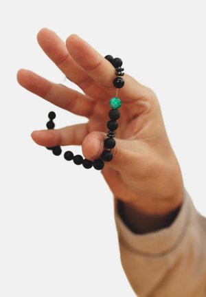 Schwarzes Perlenarmband, das in der Hand gehalten wird, bestehend aus matten und glänzenden Perlen, mit einer türkisfarbenen Perle als auffälligem Akzent.