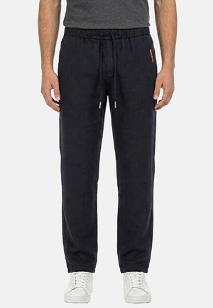 Uomo con pantaloni neri a coulisse e sneakers bianche, in piedi con le mani rilassate lungo i fianchi su sfondo semplice.