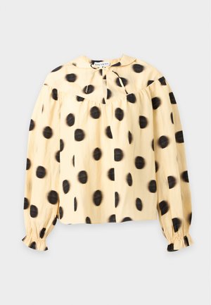 Kvinde iført en gul bluse med lange ærmer med store sorte polkaprikker og brede sorte bukser, stående med den ene hånd i lommen.