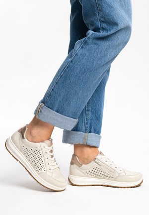 Person wearing light beige perforated sneakers with laces and side zippers, paired with rolled-up blue denim jeans.
