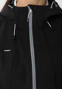 Sort vandtæt jakke med en tyk hætte, grå lynlåse, justerbare snore og et logo på brystet. Struktureret stof med stikninger som detaljer.