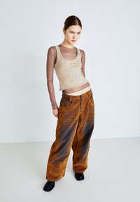 Débardeur beige avec lettrage texturé, superposé à une chemise longue à manches transparentes marron. Pantalons amples orange-brun avec un effet dégradé. Chaussures noires.