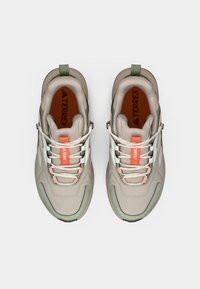 Αθλητικά παπούτσια σε ανοιχτό μπεζ με υφή στον επάνω μέρος, πράσινες λεπτομέρειες, λευκές ρίγες και πορτοκαλί λογότυπο, με κορδόνια και στιβαρή σόλα.