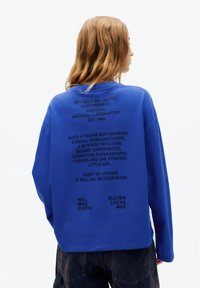 Bluza albastră cu mâneci lungi, cu text negru pe spate, incluzând detalii despre proiect și numele personajelor dintr-un serial. Material din bumbac.