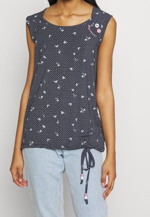 Mouwloos marineblauw shirt met witte polkadots en bloemenprint, met gevlochten koord met kralen aan de zoom, gedragen met lichtblauwe spijkerbroek.