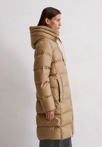 Gaisma bēša pūkainā jaka ar kapuci, horizontālu šuvēm un divām sānu kabatām. Gluda, spīdīga auduma ar siltu tekstūru.