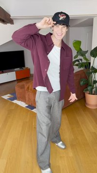 Bordová košeľa na zapínanie, biele tričko, sivé nohavice a strieborné tenisky. Čierna čiapka s vyšívaným vzorom. Vnútorné prostredie s zeleňou.