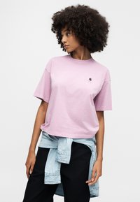 Roze katoenen T-shirt met een ronde hals en korte mouwen, voorzien van een klein logoplaatje op de borst. Draagt een licht denim jack om de taille gestrikt.