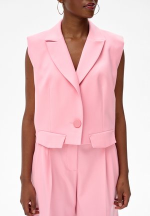 Różowy bezrękawnik z szerokimi klapami i pojedynczym guzikiem, noszony z pasującymi, wysokim stanem spodniami na osobie z kolczykiem kołowym.