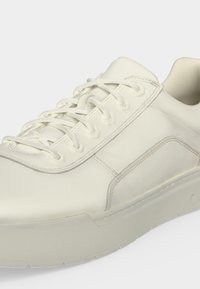 Άσπρο χαμηλό sneaker με ραφτά λεπτομέρειες, στρογγυλή μύτη, δέσιμο με κορδόνια και υφασμένο πάτο σε απλό φωτεινό φόντο.