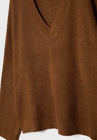 Maglione di colore marrone a maglia, con scollo a V, texture a coste e una vestibilità ampia. L'orlo è leggermente arrotondato con spacchetti laterali per facilitare i movimenti.