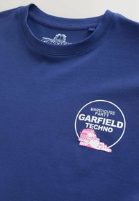 T-shirt bleu avec un dessin animé de Garfield rose allongé, texte "Warehouse Party Garfield Techno" à l'intérieur d'un cercle blanc sur la poitrine.