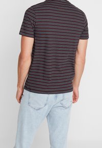 Camisa de manga corta para hombre con rayas horizontales en negro, gris y rojo. Material de algodón, cuello redondo, diseño ligeramente ajustado. Jeans de mezclilla clara.