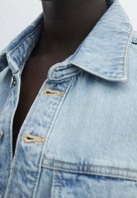 Veste en denim bleu clair avec col classique, fermeture à boutons sur le devant avec coutures visibles et finition texturée.