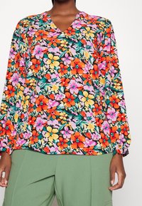 Donna che indossa una blusa a maniche lunghe con scollatura a V, caratterizzata da una vivace stampa floreale arancione, gialla e rosa, abbinata a pantaloni verdi.