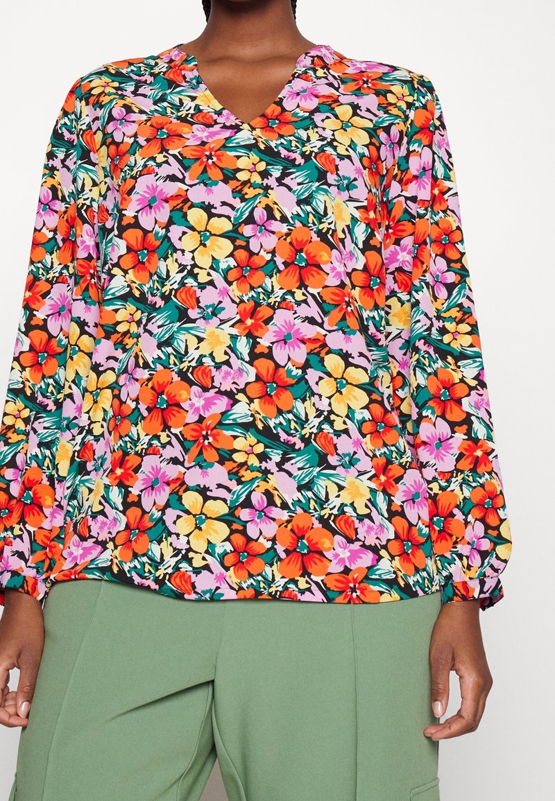 Donna che indossa una blusa a maniche lunghe con scollatura a V, caratterizzata da una vivace stampa floreale arancione, gialla e rosa, abbinata a pantaloni verdi.