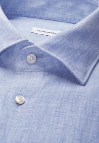 Chemise en lin bleu clair avec col pointu et boutons blancs, avec étiquette de marque visible à l'intérieur du col.