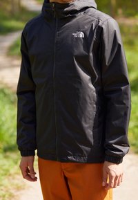 Svart vattentät jacka med frontdragkedja, elastiska ärmar och huva, med en logo på bröstet. Bärs över orange byxor.