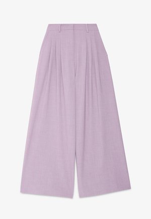 Vrouw staand in een eenvoudige kamer, gekleed in een lila broekpak met wijde pijpen, een wit topje, bruine schoenen en oorbellen in de vorm van ringen.