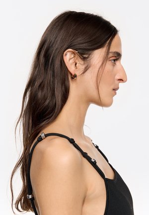 Vêtement sans manches noir avec des bretelles nouées, présentant des accents en métal argenté. Le mannequin montre un profil de côté avec de longs cheveux et des boucles d'oreilles créoles.