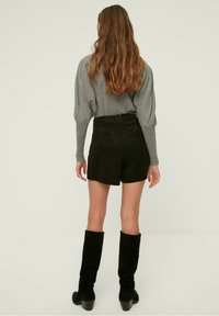 Shorts en daim noir à taille haute, associés à un pull gris à manches longues. Modèle portant des bottes noires montantes. Fond clair.