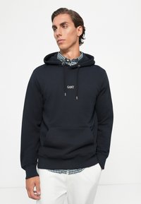 Joven vestido con una sudadera azul marino sobre una camisa de cuadros verde y blanca, combinada con pantalones blancos, de pie frente a un fondo blanco liso.