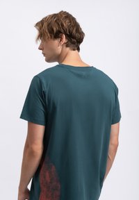 Teal katoenen T-shirt met korte mouwen, voorzien van een abstract rood ontwerp rechtsonder en een logo-detail bij de achterste halslijn.