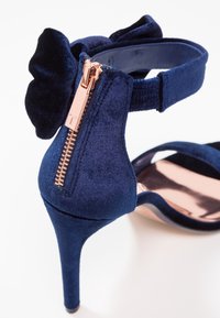 Mörkblå sammet högklackad sandal med dragkedja bak, blommig dekoration och texturerade remmar.