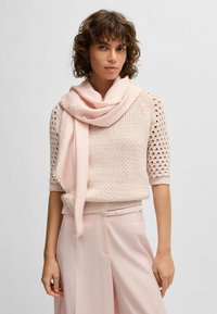 Maglione a maglia rosa chiaro con maniche corte caratterizzato da una texture a motivo. Sciarpa rosa drappeggiata intorno al collo, abbinata a pantaloni rosa chiaro.