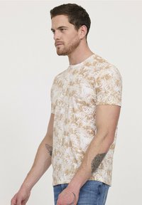 T-shirt à manches courtes en blanc avec un motif de feuille de palmier beige. Le tissu en coton offre une texture lisse. Design à col rond, style ajusté.