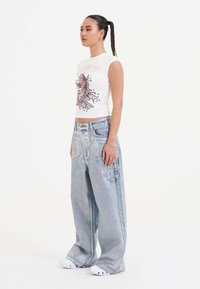 White sleeveless top with pink graphic, paired with loose, light blue denim jeans featuring embroidered accents. Model stands in profile.