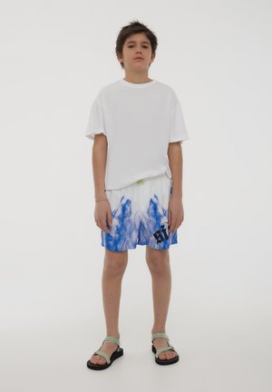 T-shirt bianco oversize, pantaloni corta tie-dye blu e bianchi, sandali verdi con cinture regolabili, in piedi contro uno sfondo bianco semplice.