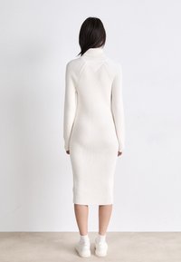 Vestido de punto de manga larga en color crema, con textura acanalada, silueta ajustada y cuello alto, combinado con zapatillas blancas y calcetines.