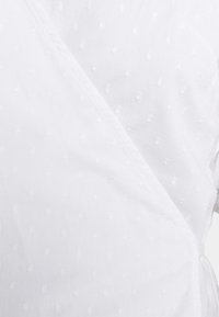 Blouse blanche et légère avec un design cache-cœur, présentant des points texturés et une finition lisse. Les détails des coutures sont visibles le long des bords.