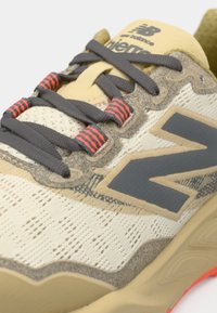 Chaussure de course beige avec empeigne en maille texturée, accents gris foncé et lacets rayés rouges. Dispose d'une semelle coussinée à la forme distinctive.