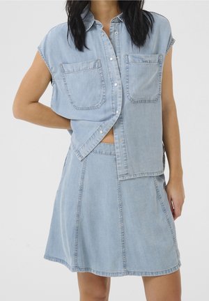 Femme portant une chemise en denim bleu clair sans manches avec des poches avant et une jupe en denim assortie sur un fond uni.