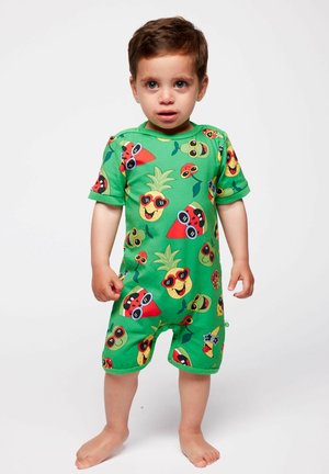 Tout-petit debout pieds nus portant une combinaison verte avec des personnages de fruits colorés portant des lunettes de soleil sur un fond blanc.