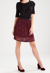 Haut noir avec un col froncé, associé à une jupe en dentelle bordeaux avec un motif floral. Porte des chaussures à talons hauts bordeaux.