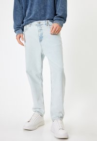Jeans de denim azul claro con un corte relajado, que cuentan con un cierre de botón en la parte delantera y un diseño clásico de cinco bolsillos. Llevados con zapatillas blancas.
