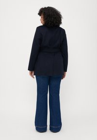 Marineblazer mit tailliertem Schnitt und Gürtel in der Taille. Kombiniert mit lässigen, weit geschnittenen blauen Jeans, die einen entspannten, strukturierten Stil zeigen.