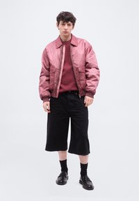 Νέος άνδρας που φοράει ροζ σατέν μπουφάν τύπου μπομπέρ, μπορντό πλεκτό πουλόβερ με υφή, μαύρες σορτς φαρδιάς γραμμής, μαύρες κάλτσες και μαύρα δερμάτινα μοκασίνια.