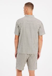 Conjunto de camisa de manga corta y pantalones cortos, hecho de tela a rayas en beige y negro. Presenta un collar y un corte relajado, con detalle de bolsillo en los pantalones cortos.