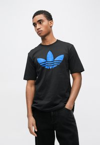 Melna kokvilnas T-krekls ar zilā Adidas logotipu un trim svītrām, īsām piedurknēm un apaļu kakla izgriezumu. Gluda tekstūra un standarta piegriezums.