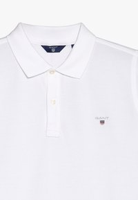 Valkoinen polo-paita, jossa lyhyt nappilista, viistopainikkeinen kaulus ja pieni GANT-logo brodeerattuna vasempaan rintaan.