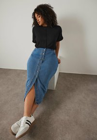 Jupe crayon en denim avec des boutons sur le devant et une fente sur le côté, associée à un haut en tricot noir à côtes et des baskets blanches avec des rayures noires.