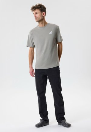 Homme portant un t-shirt gris avec un petit motif d'agrumes, un pantalon noir et des sabots noirs, debout et regardant vers la gauche sur un fond blanc uni.