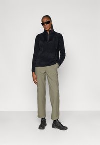 Sort fleece trøje med halv lynlås, parret med khaki vide bukser og sorte sneakers. Model med sorte solbriller.