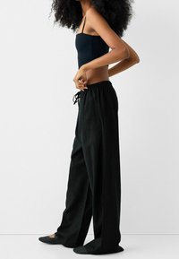 Pantalons larges noirs avec une taille élastique et un cordon de serrage. Associés à un débardeur noir ajusté. Portés avec des ballerines noires texturées.
